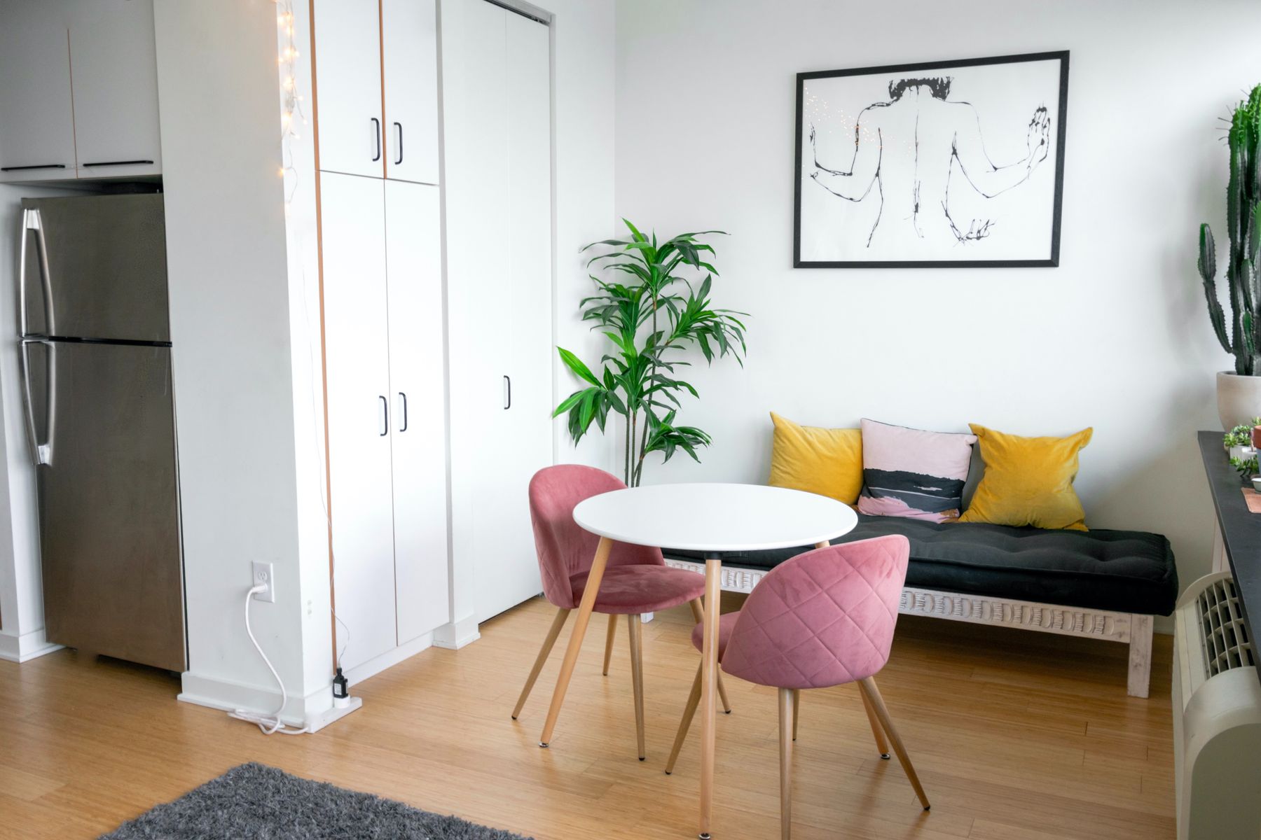 Petit studio lumineux avec table ronde, chaises roses, banquette à coussins jaunes et plantes vertes